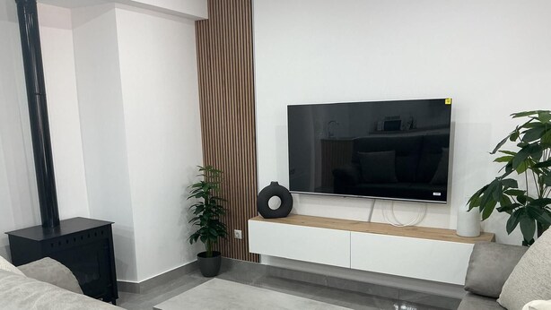 Un salón moderno con una televisión de pantalla plana montada en un mueble blanco, sofás grises, una mesita de noche negra y una planta en maceta.