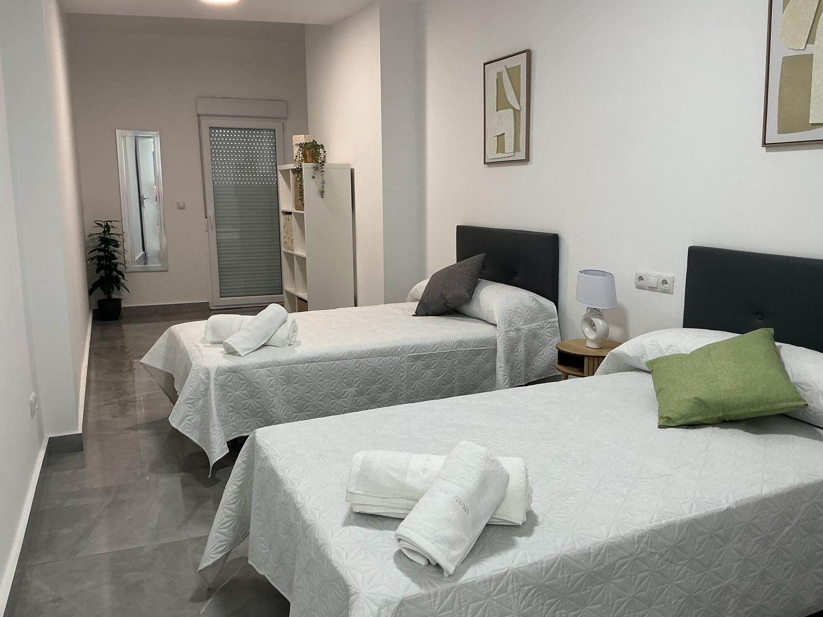 Habitación de hotel con dos camas, una mesita de noche con lámpara, una planta en maceta y una puerta con ventanal.