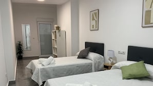 Habitación de hotel con dos camas, una mesita de noche con lámpara, una planta en maceta y una puerta con ventanal.