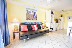 A living room with a black leather sofa, two lamps, a painting, and a kitchen in the background. - 4201 Coastal Highway Unit 404 (Ocean City)