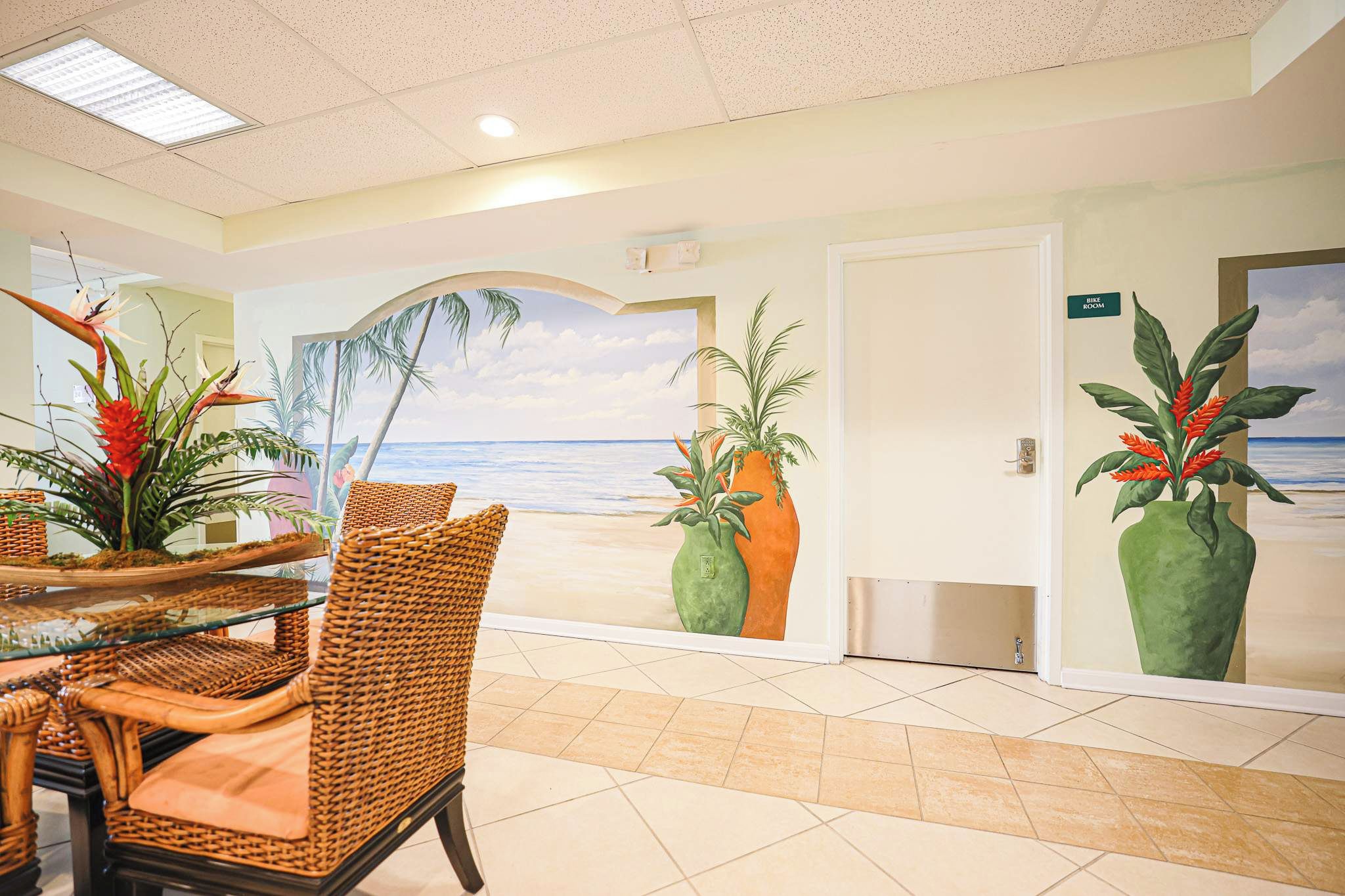 A hotel lobby with a beach view mural, wicker furniture, and potted plants.