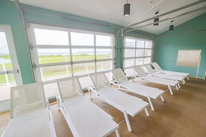 A room with multiple white sunbeds arranged in rows, large windows with a view of the outdoors, and a green wall. - 4201 Coastal Highway Unit 404 (Ocean City)