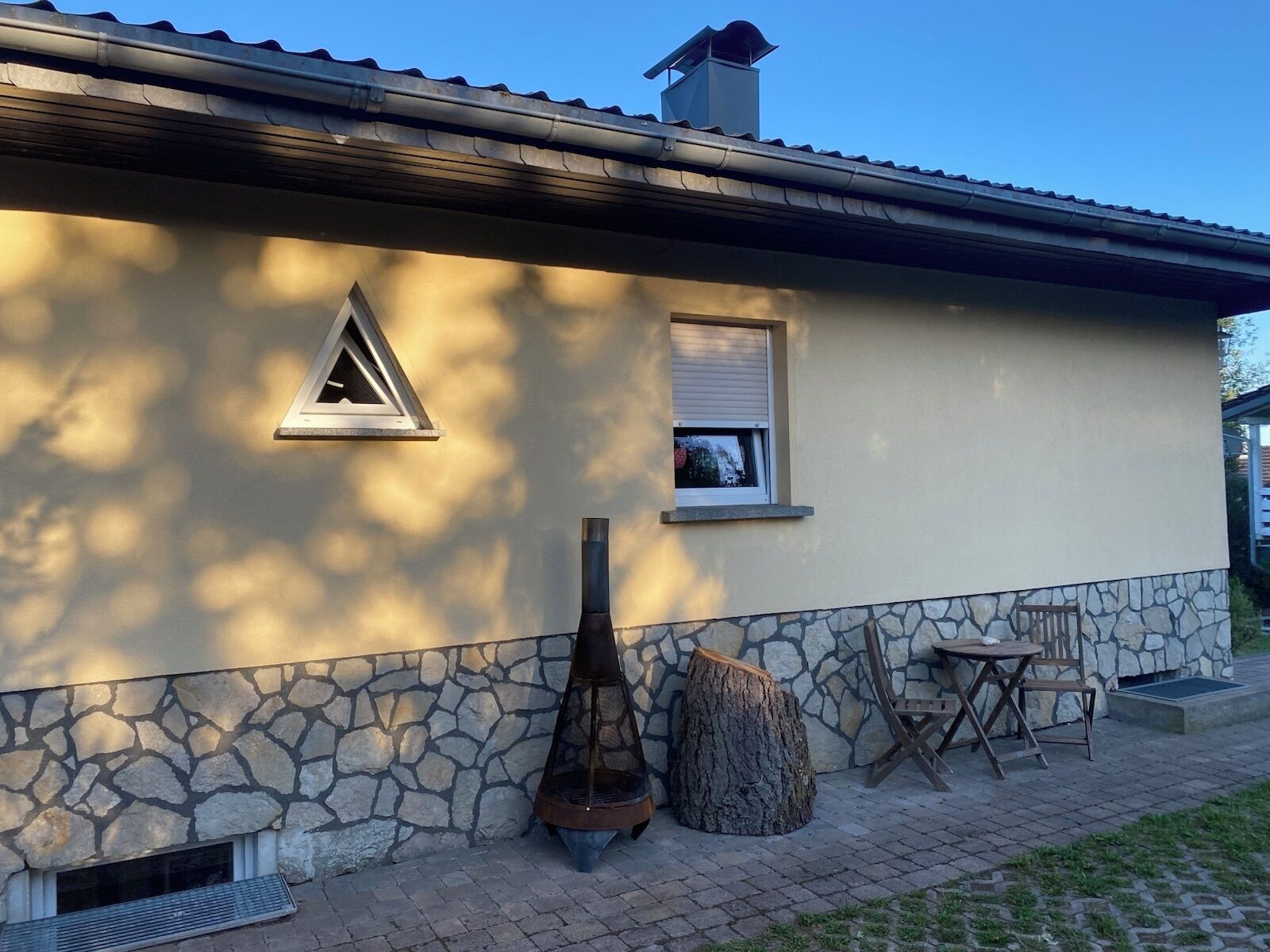 Un edificio de fachada de piedra, con chimenea, ventanal triangular y una mesa de madera con sillas.