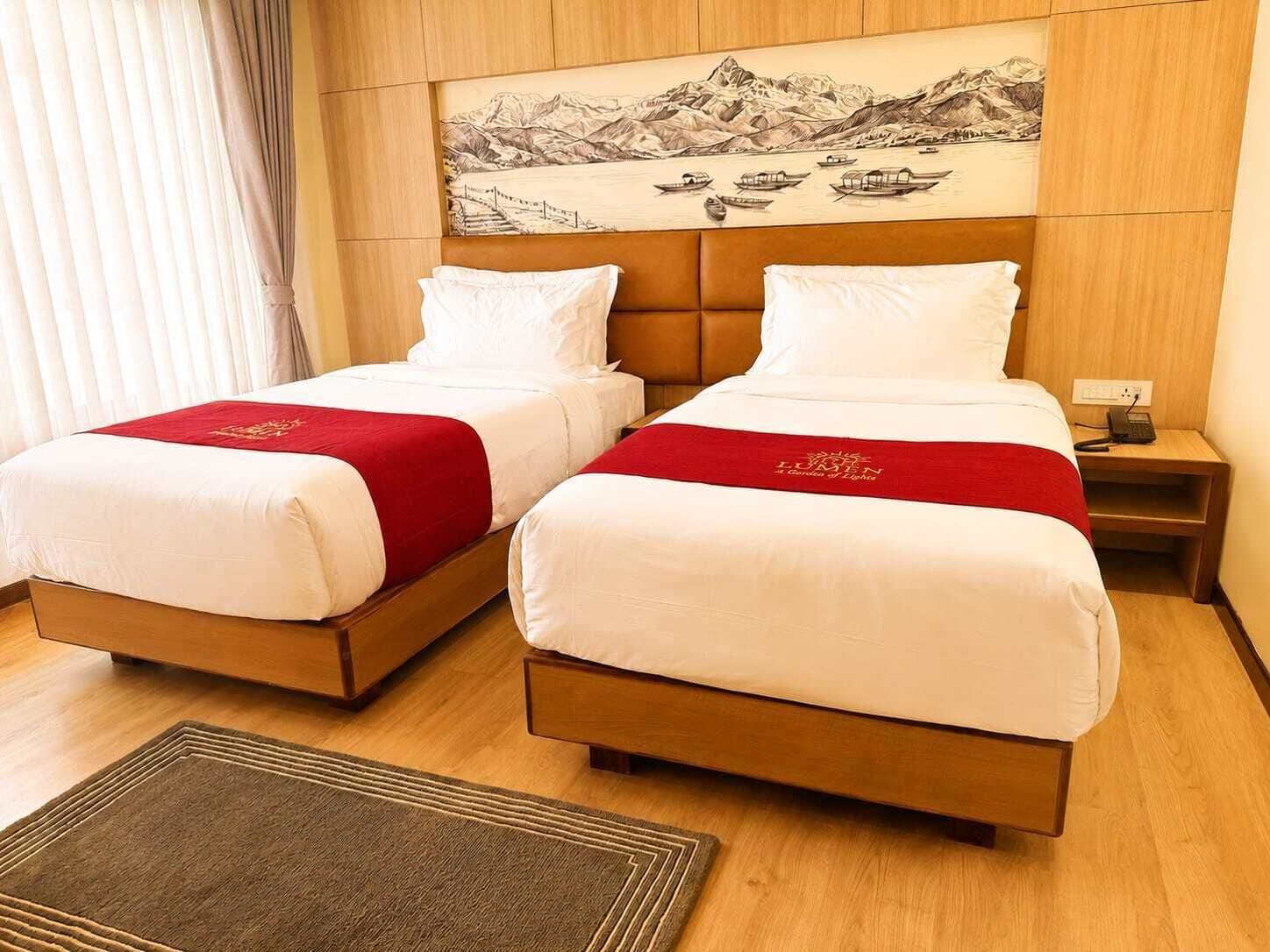 Habitación de hotel con dos camas, cabecero de madera, mesita de noche y un cuadro de un paisaje montañoso.