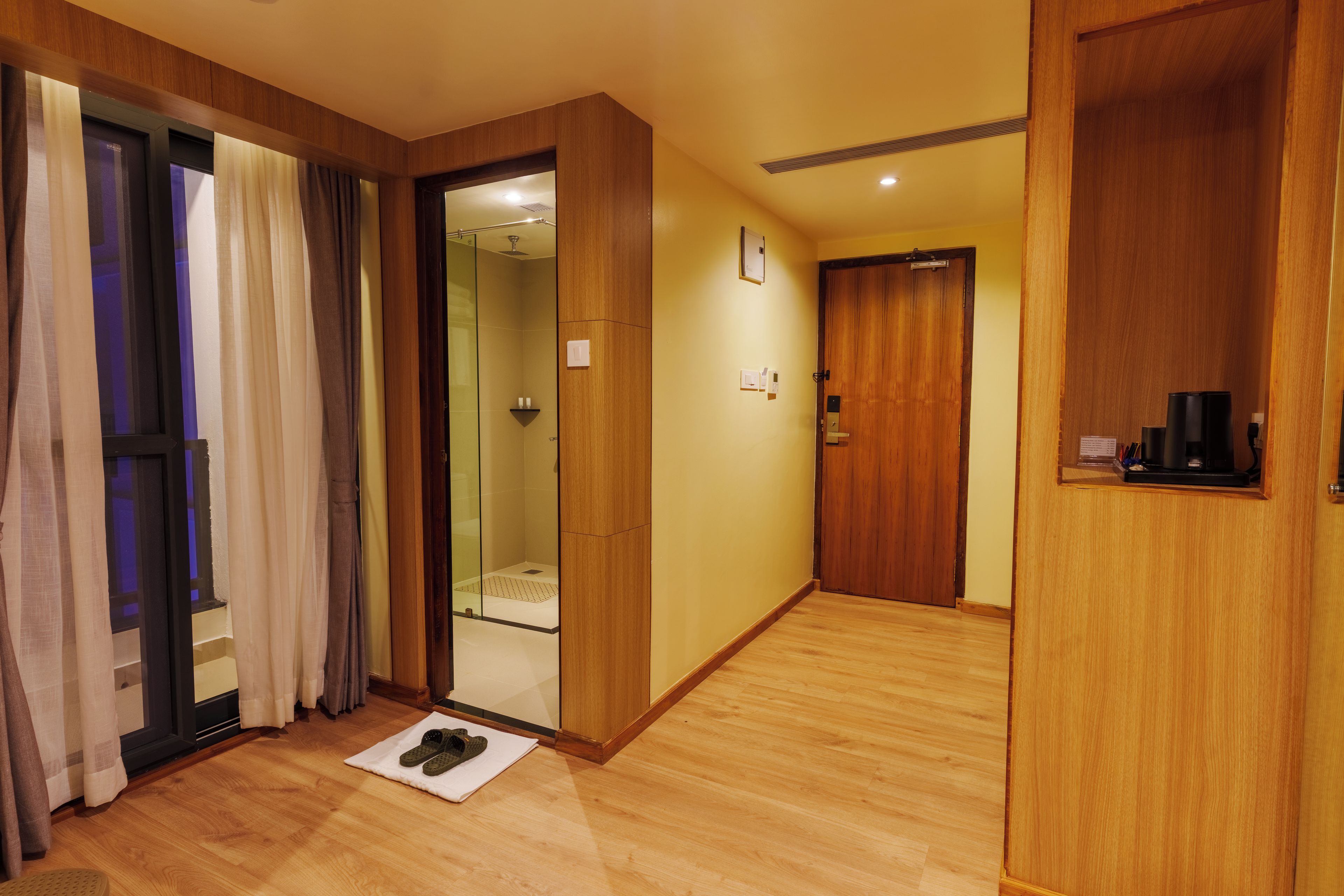 Una camera d'albergo con pavimento in legno, box doccia in vetro, armadio in legno e un piccolo tappetino con ciabatte.