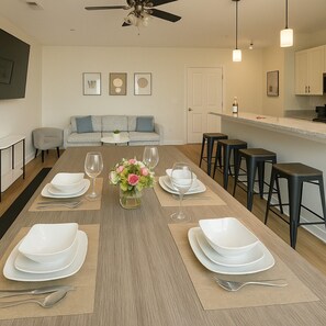 A modern dining area with a long table set for four, a sofa in the background, and a kitchen area with bar stools. - Family & Group Retreat3QueensBeds + Pool Table 7B (Birmingham)