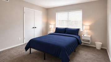 A bedroom with a bed, two bedside tables, a window with blinds, a door, and a wall vent.