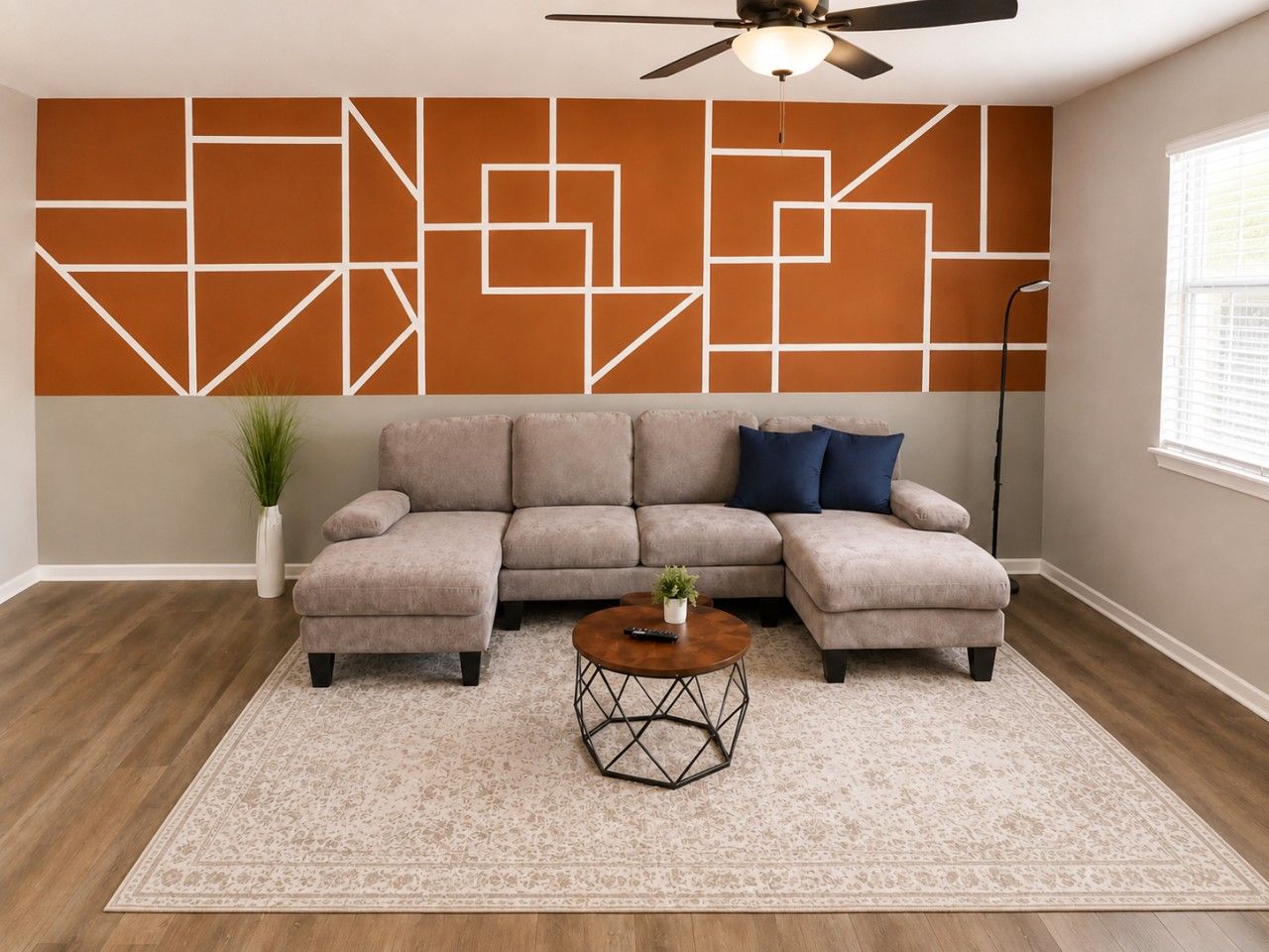 A living room with a sectional sofa, a coffee table, a wall with geometric patterns, a ceiling fan, and a window with blinds.