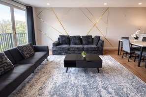 Ein modernes Wohnzimmer mit einem grauen Sofa, einem schwarzen Couchtisch und einem Essbereich mit weißem Tisch und Stühlen.