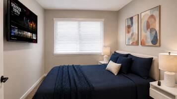 Ein Schlafzimmer mit einem Bett, einem an der Wand befestigten Fernseher, einem Fenster mit Jalousien und zwei Wandleuchten.