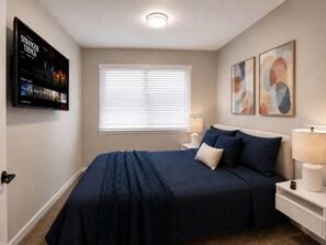 A bedroom with a bed, a TV mounted on the wall, a window with blinds, and two wall-mounted lamps.