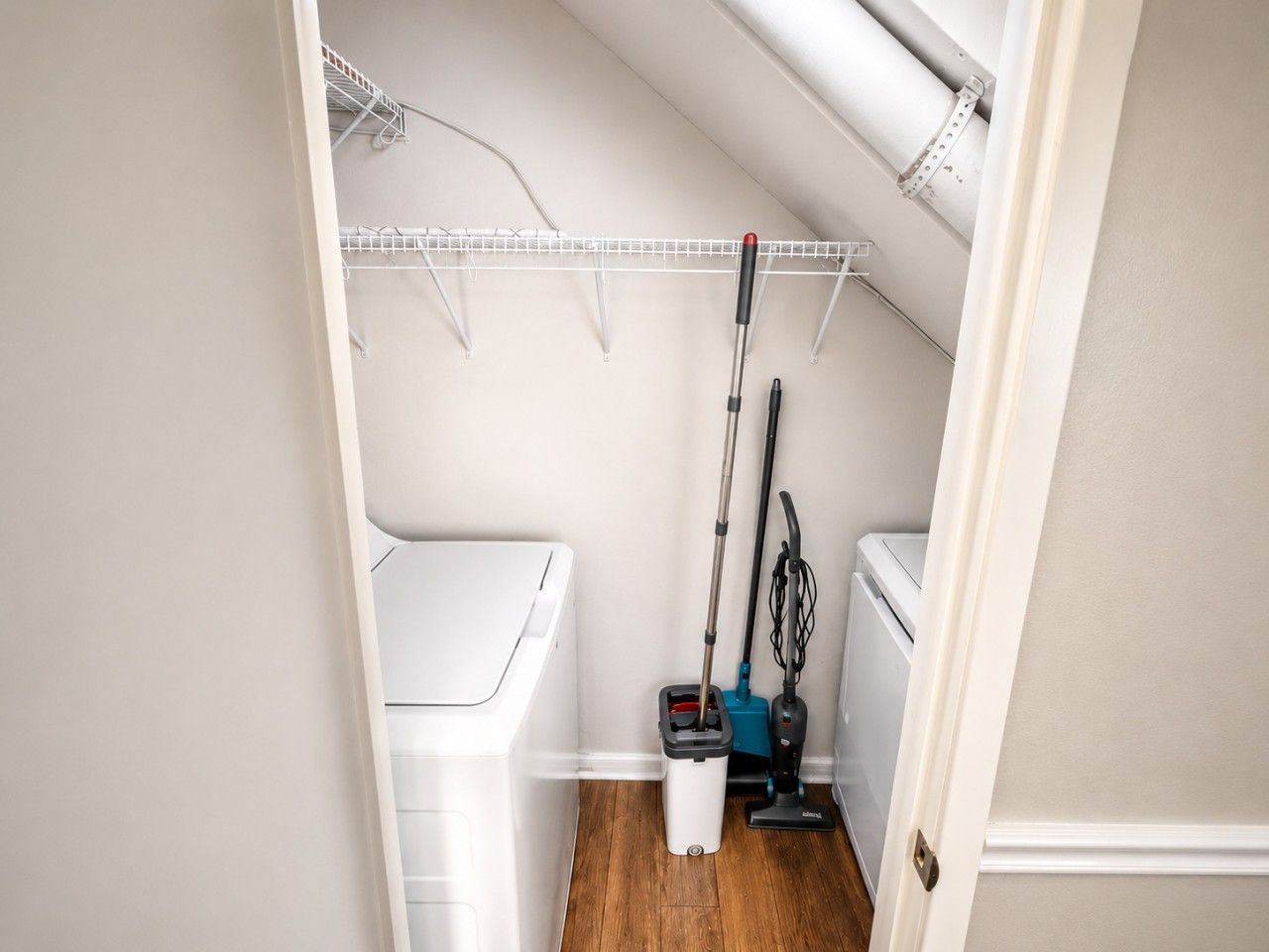 A small laundry room with a white washing machine, a blue and black vacuum cleaner, and a mop on the floor.