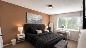 A bedroom with a bed, bedside tables, a desk, a chair, a window with blinds, and a painting on the wall. - Peaceful 2BR Townhome Near Birmingham (Hoover)