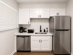 A compact kitchen with white cabinetry, a stainless steel refrigerator, a dishwasher, and a coffee maker.