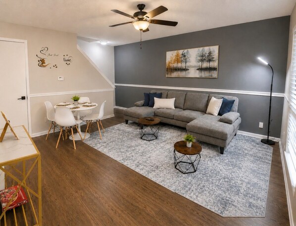A living room with a grey sofa, two coffee tables, a dining table with chairs, a ceiling fan, and a painting on the wall.