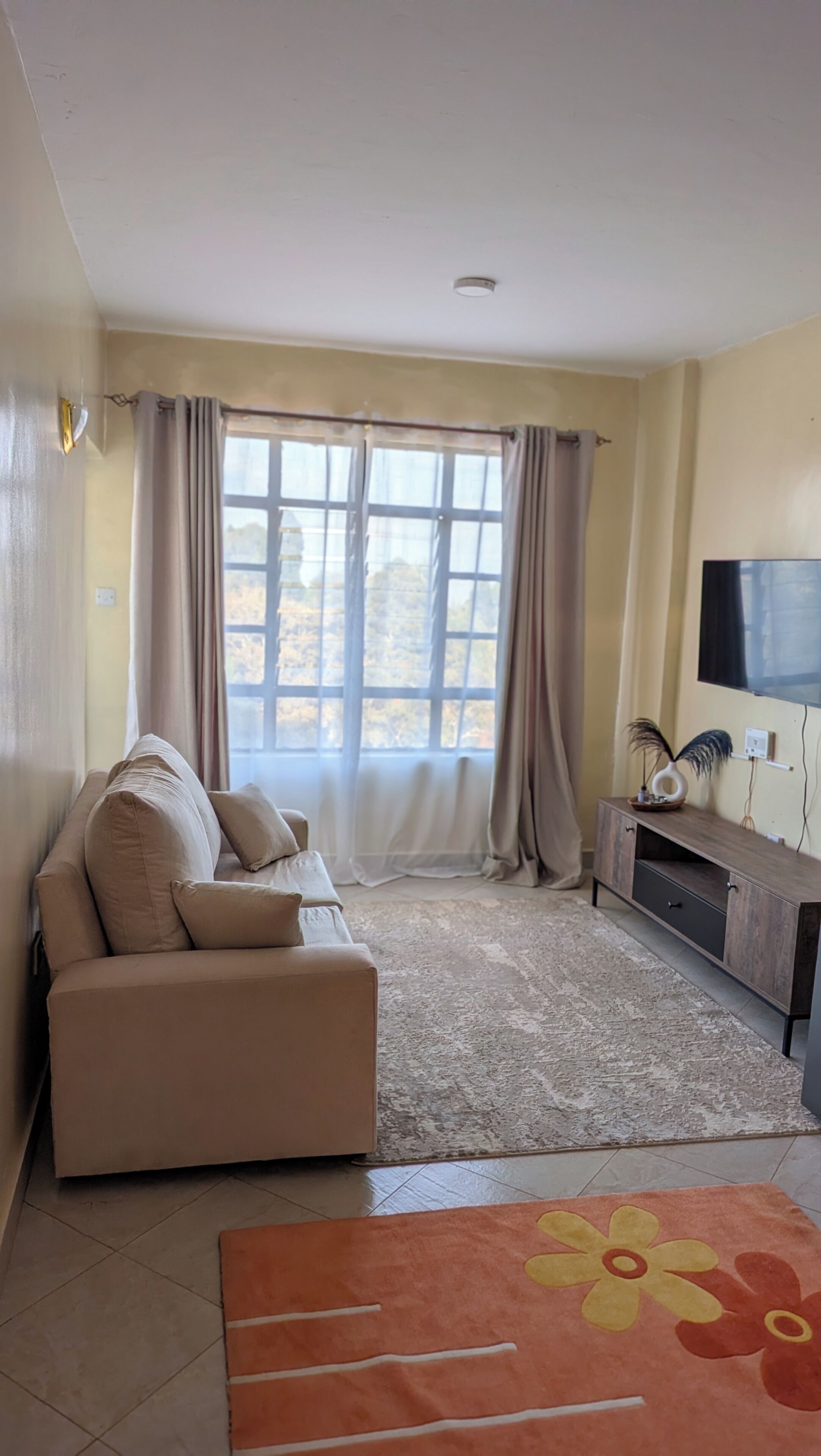 Uma sala de estar com sofá, televisão na parede e janela com cortinas.