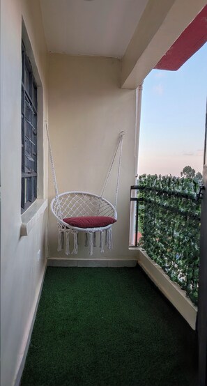 A balcony with a macrame swing chair, a window with metal bars, and a green artificial turf floor.