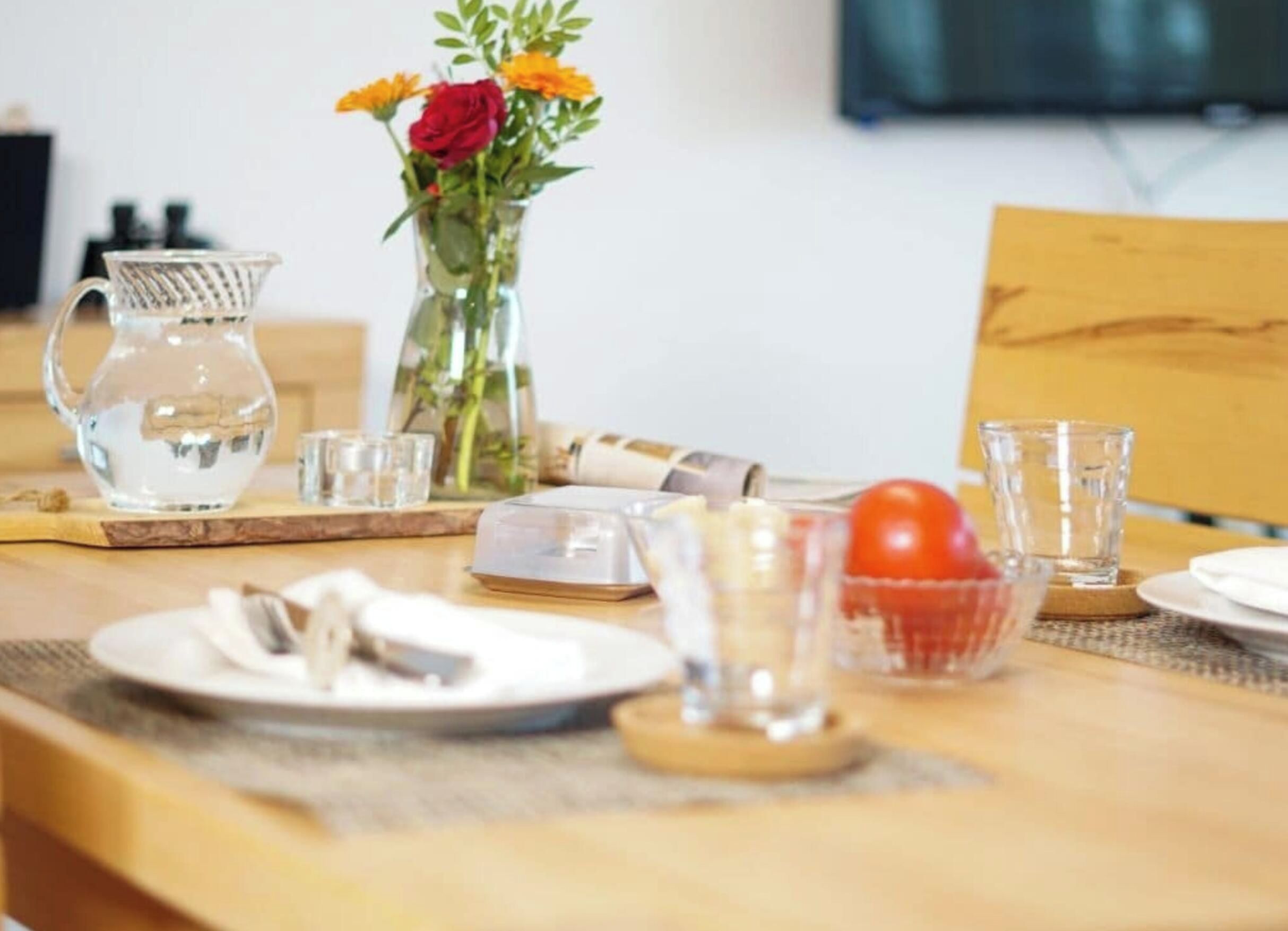Una mesa de comedor dispuesta con platos, vasos, una jarrón y un cuenco con tomate.