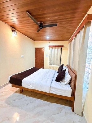 Un dormitorio con techo de madera, una cama con ropa de cama blanca y oscura, un ventilador de techo, una ventana con cortinas y una puerta.