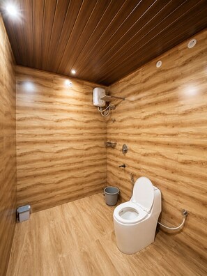 A compact bathroom with wooden walls and a sloped ceiling, featuring a toilet, a shower unit, and a small trash bin. - Venkataramana KV (Mekeri)