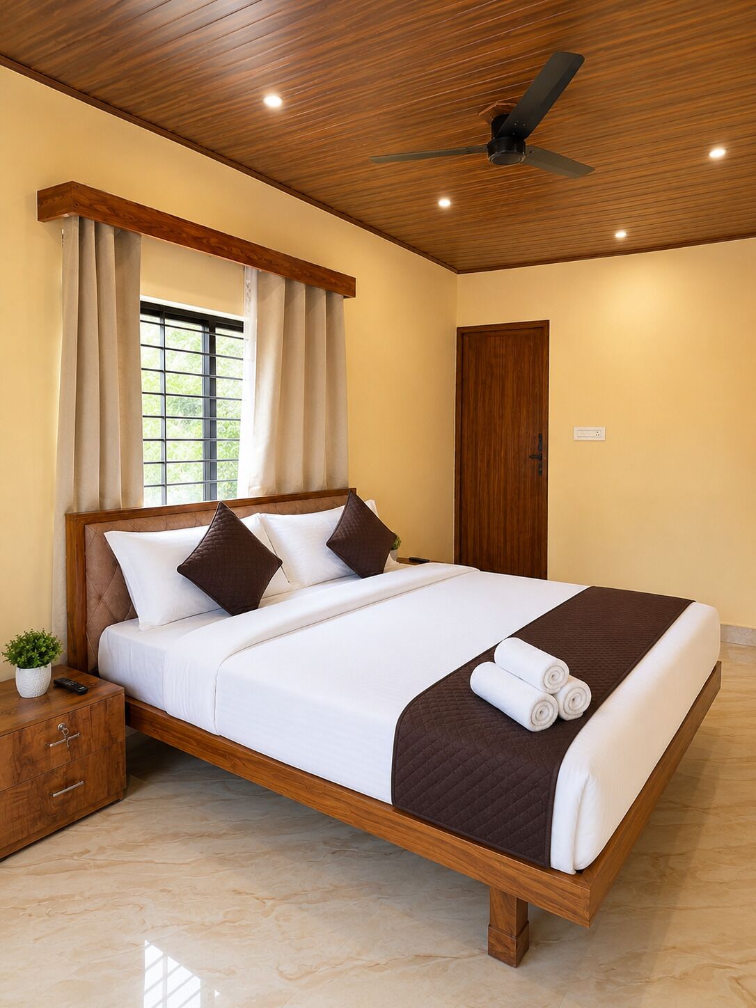 Une chambre d’hôtel avec un lit en bois, un ventilateur de plafond, une fenêtre avec des rideaux et une table de chevet en bois ornée d’une plante.