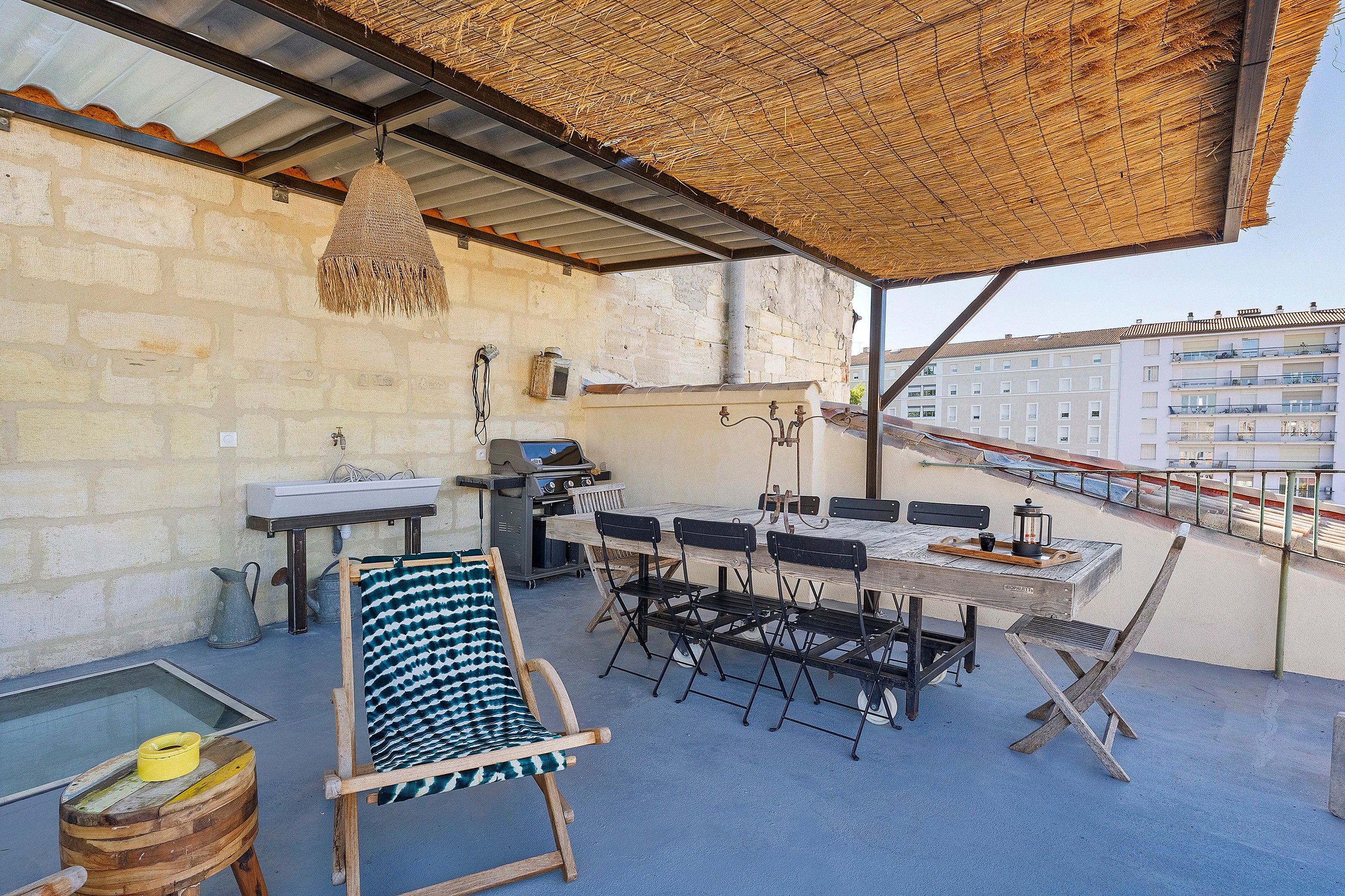 A rooftop terrace with a grill, dining table, chairs, and a hanging light fixture.