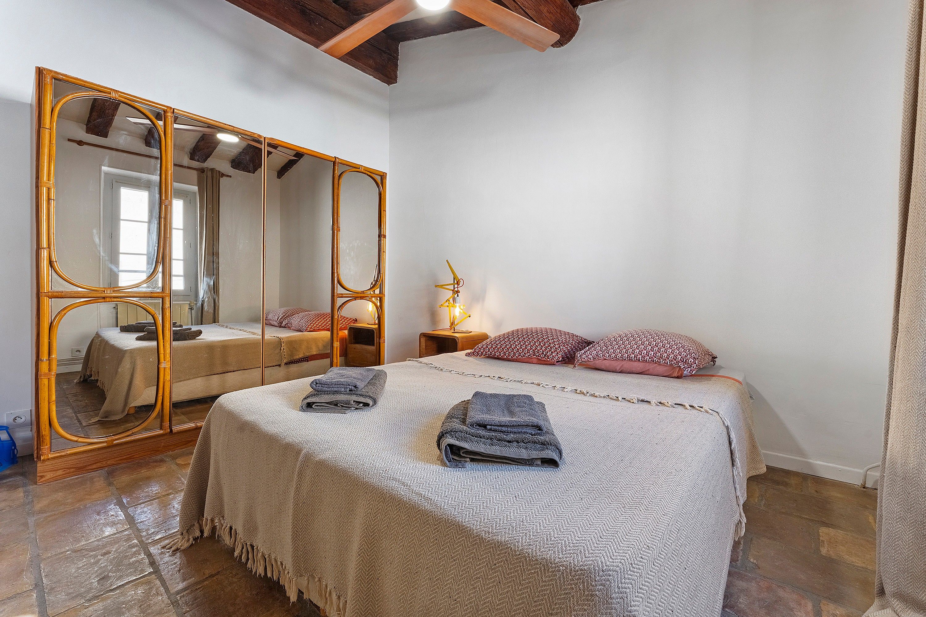 A bedroom with a large mirror, a bed with multiple pillows, a bedside table with a lamp, and a wooden floor.