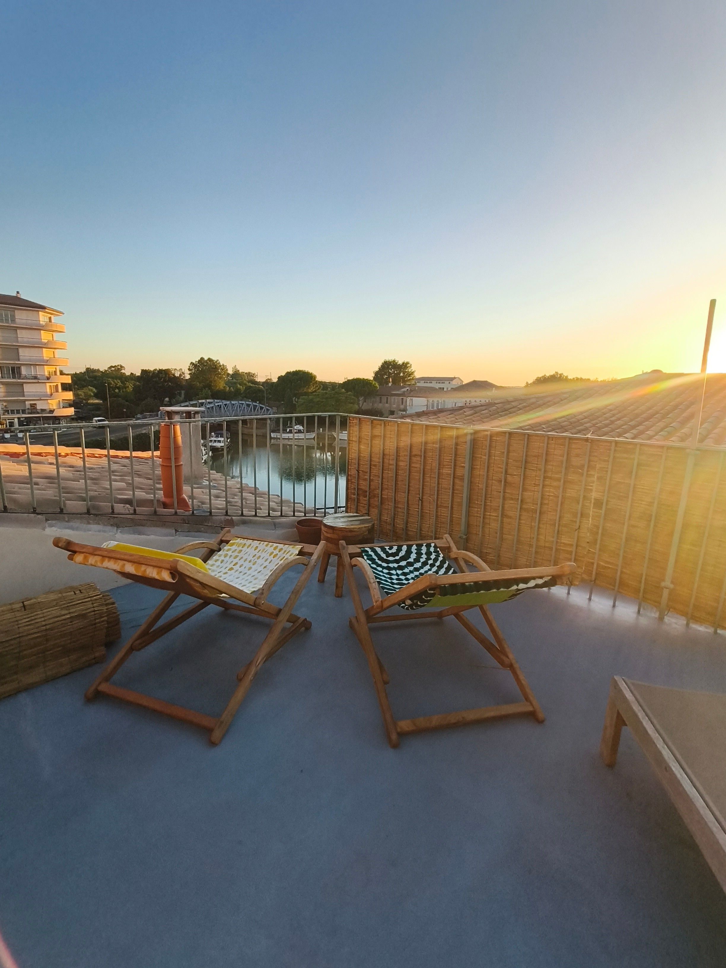 Un balcon avec deux fauteuils de jardin en bois, une vue sur une étendue d’eau et un coucher de soleil.