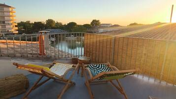 Un balcon avec deux fauteuils de jardin en bois, une vue sur une étendue d’eau et un coucher de soleil.