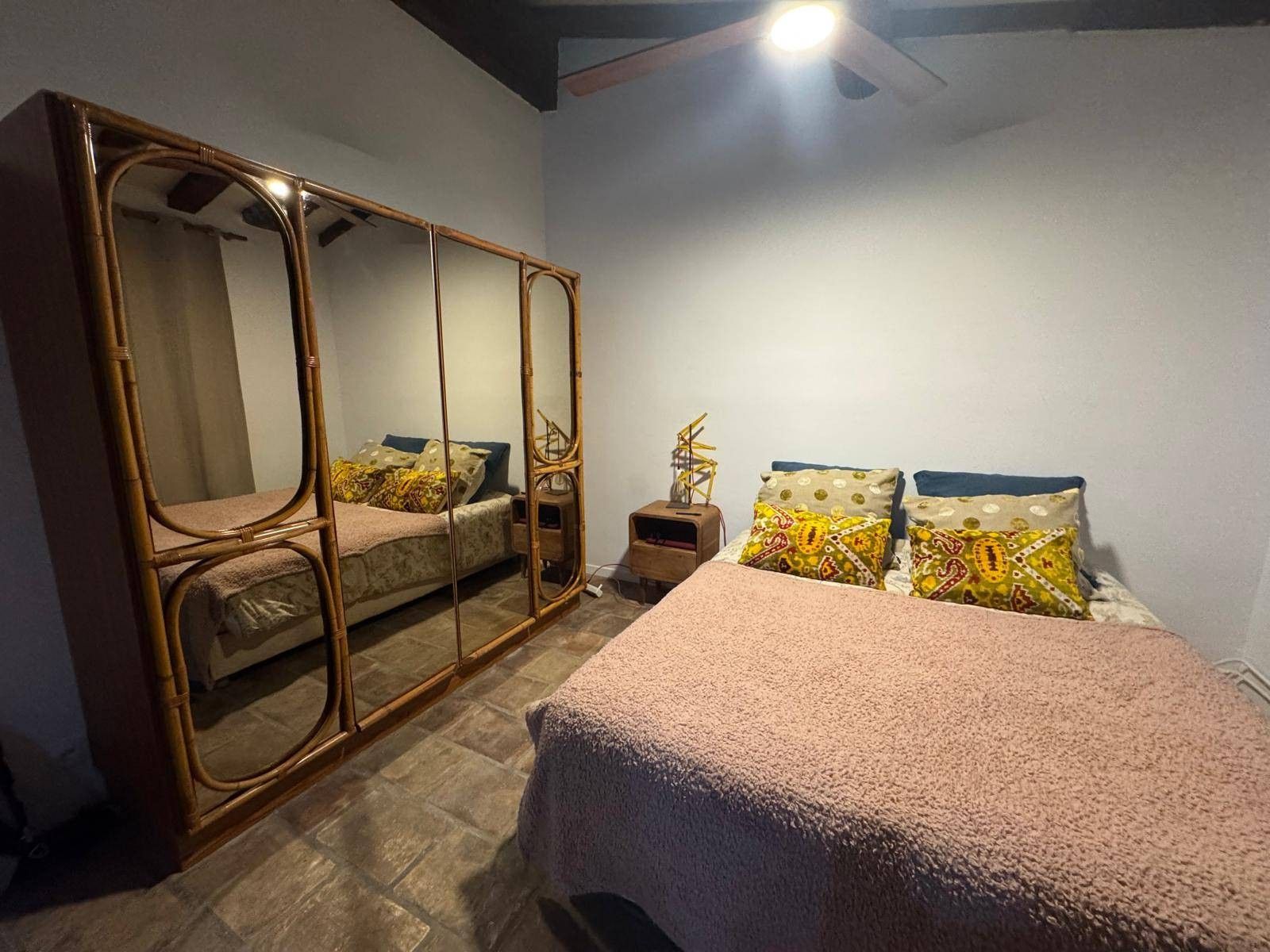 Een slaapkamer met een bed, een grote spiegel, een nachtkastje en een plafondventilator.