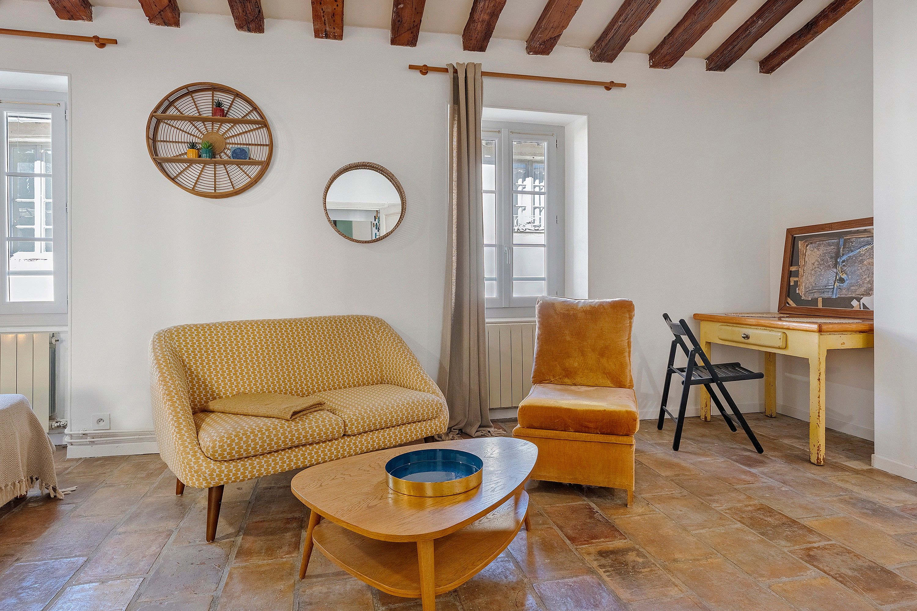 Una habitación con sofá, una silla, una mesita, un espejo y una ventana con cortinas.