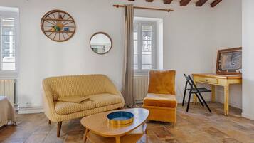 Una habitación con sofá, una silla, una mesita, un espejo y una ventana con cortinas.
