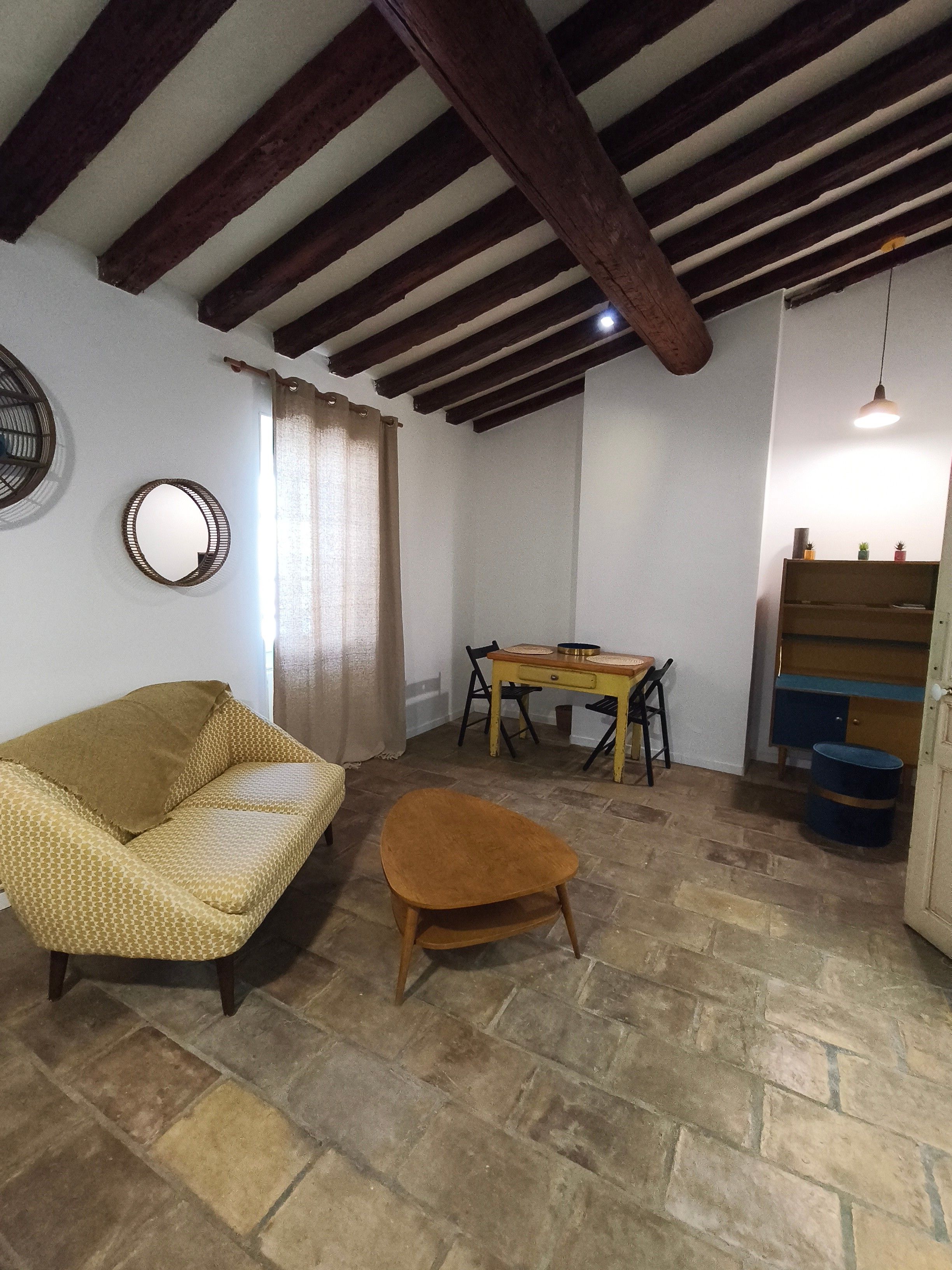 Une pièce comprenant un canapé, une petite table en bois, une chaise, un miroir et un plafond avec des poutres apparentes.