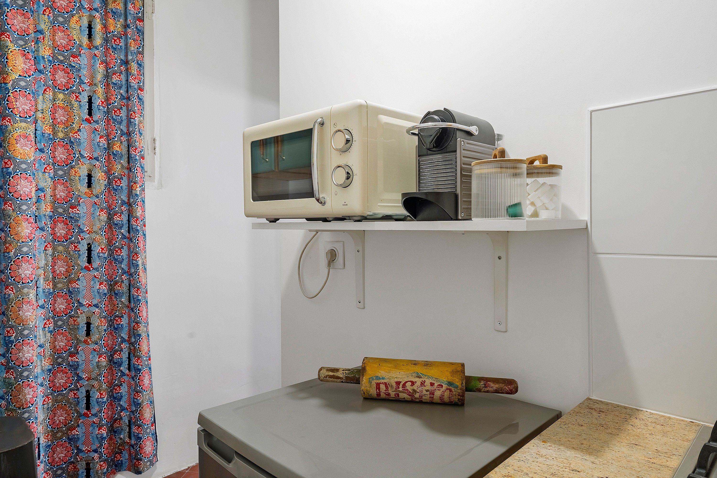 A microwave on a shelf, a small trash bin, and a patterned curtain.