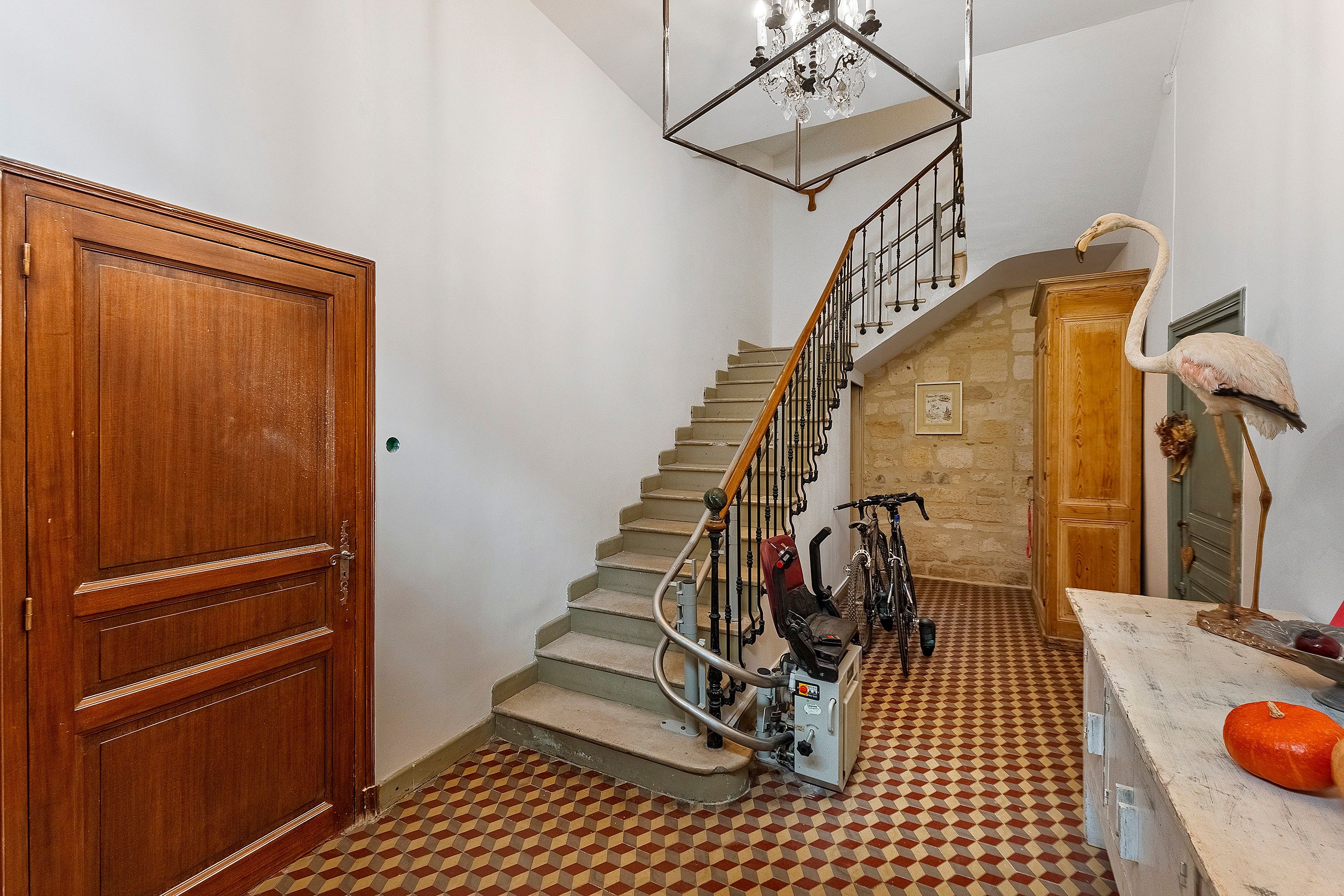 A staircase with a checkered floor, a wooden door, a chandelier, and a flamingo statue.