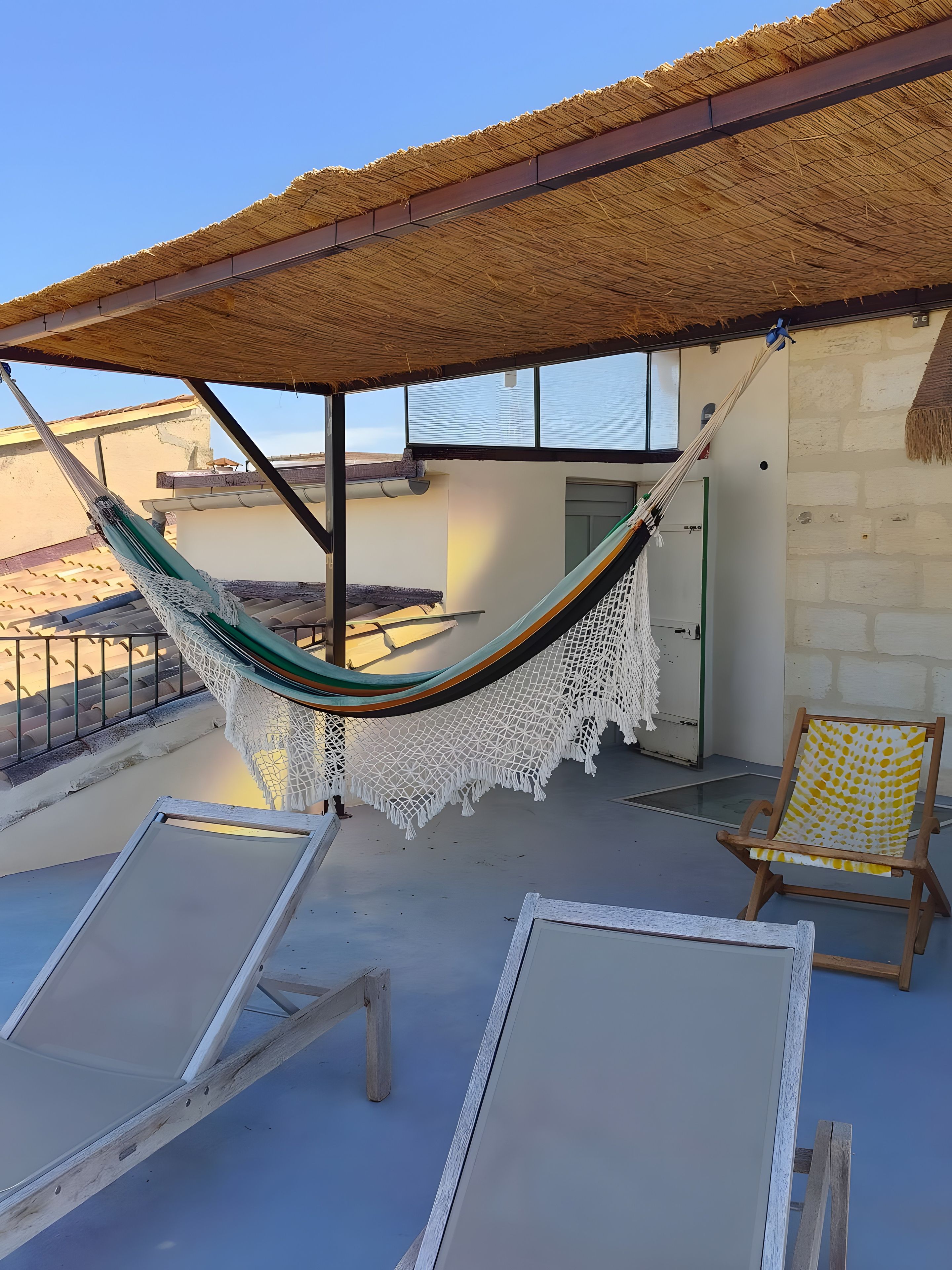 Una hamaca bajo techo de paja, dos sillones de descanso y una silla estampada en una terraza.