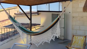 Una hamaca bajo techo de paja, dos sillones de descanso y una silla estampada en una terraza.