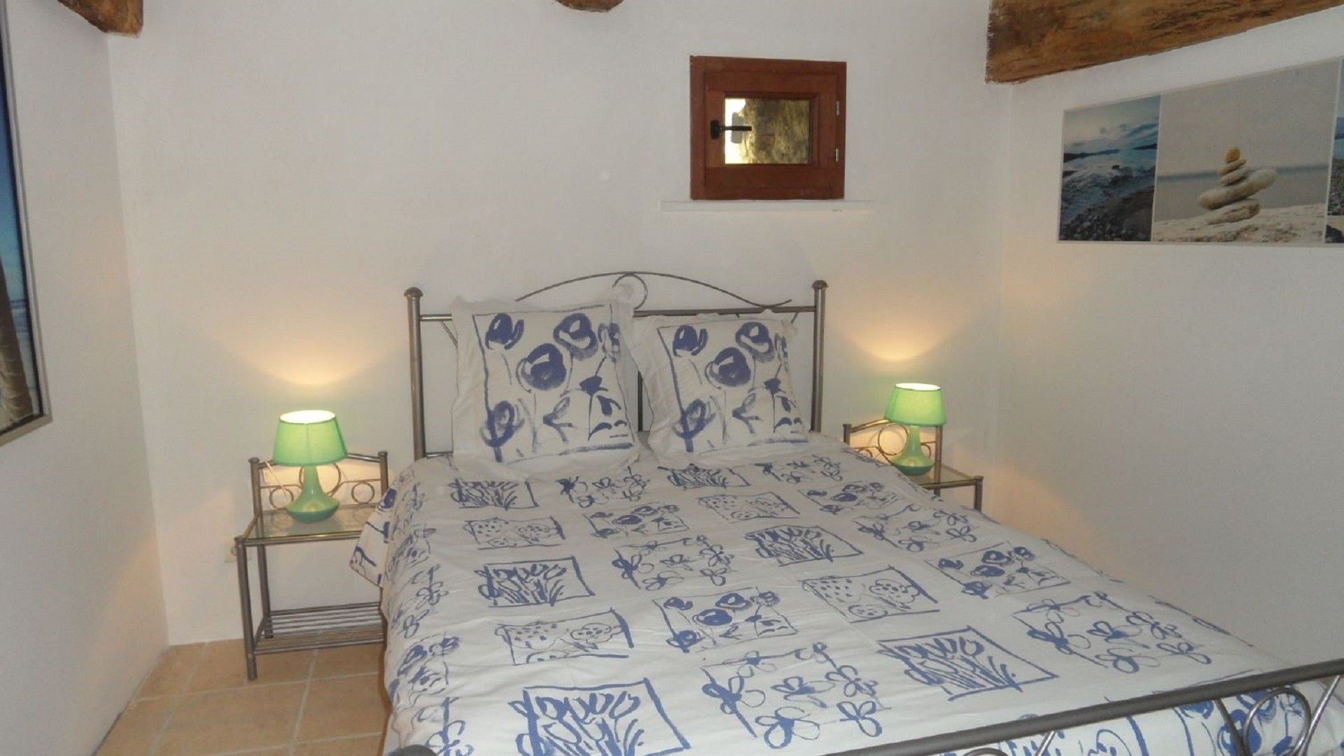 A bedroom with a bed, two bedside lamps, a framed picture on the wall, and a tiled floor.