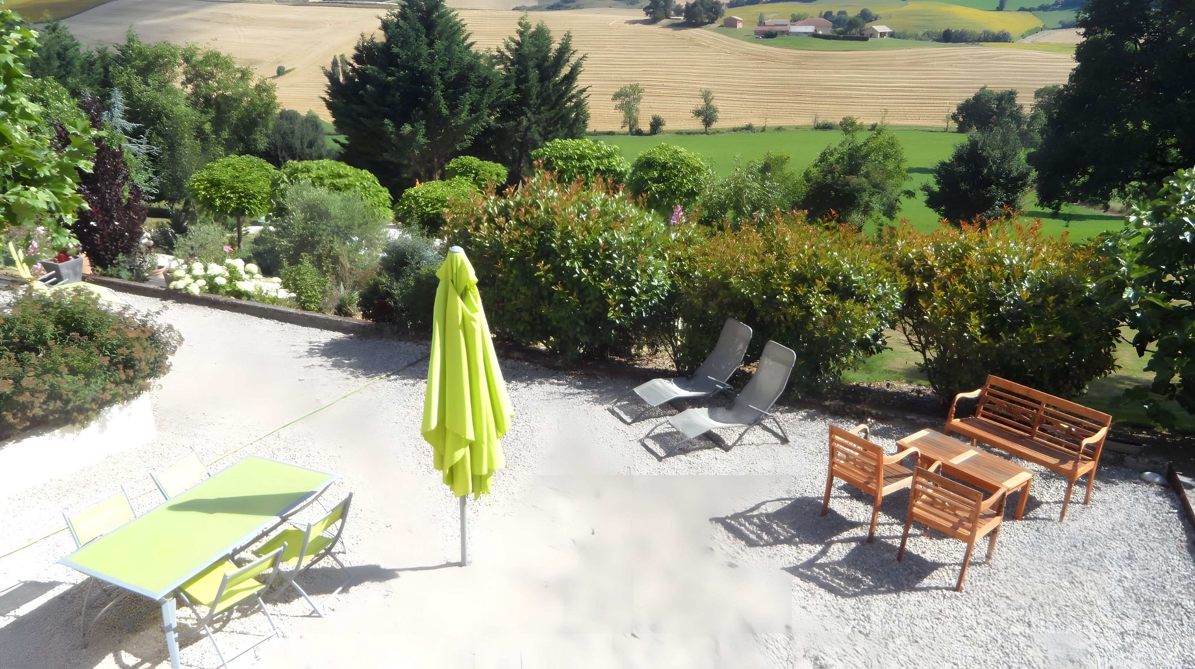 An outdoor seating area with a table, chairs, and an umbrella, overlooking a vineyard and green fields.