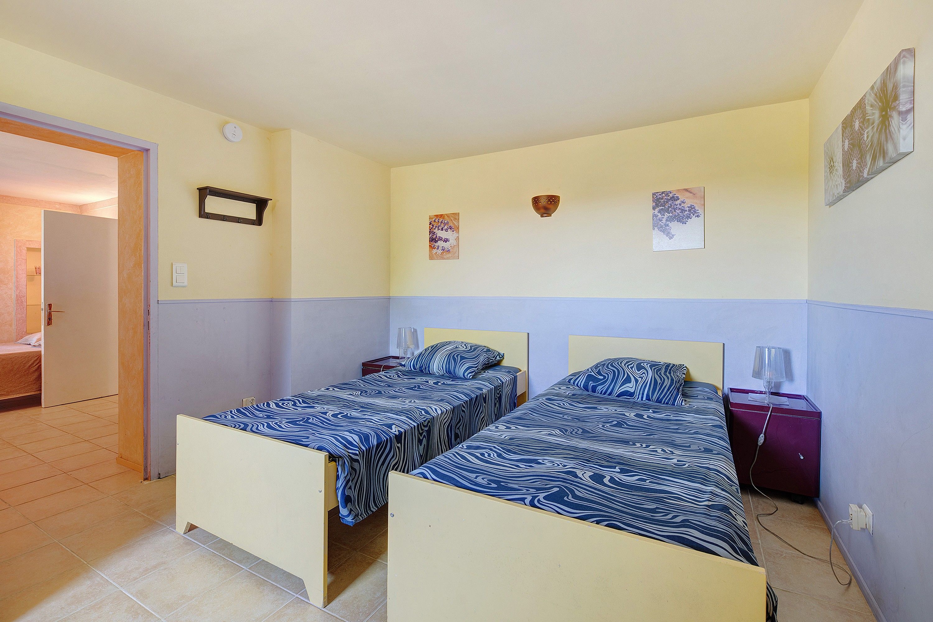A room with two beds, a door, a wall-mounted light fixture, and a small table with a lamp.