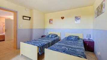 A room with two beds, a door, a wall-mounted light fixture, and a small table with a lamp.