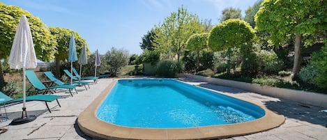 Une piscine rectangulaire avec une terrasse carrelée, entourée d’arbres verdoyants et de chaises longues bleues.