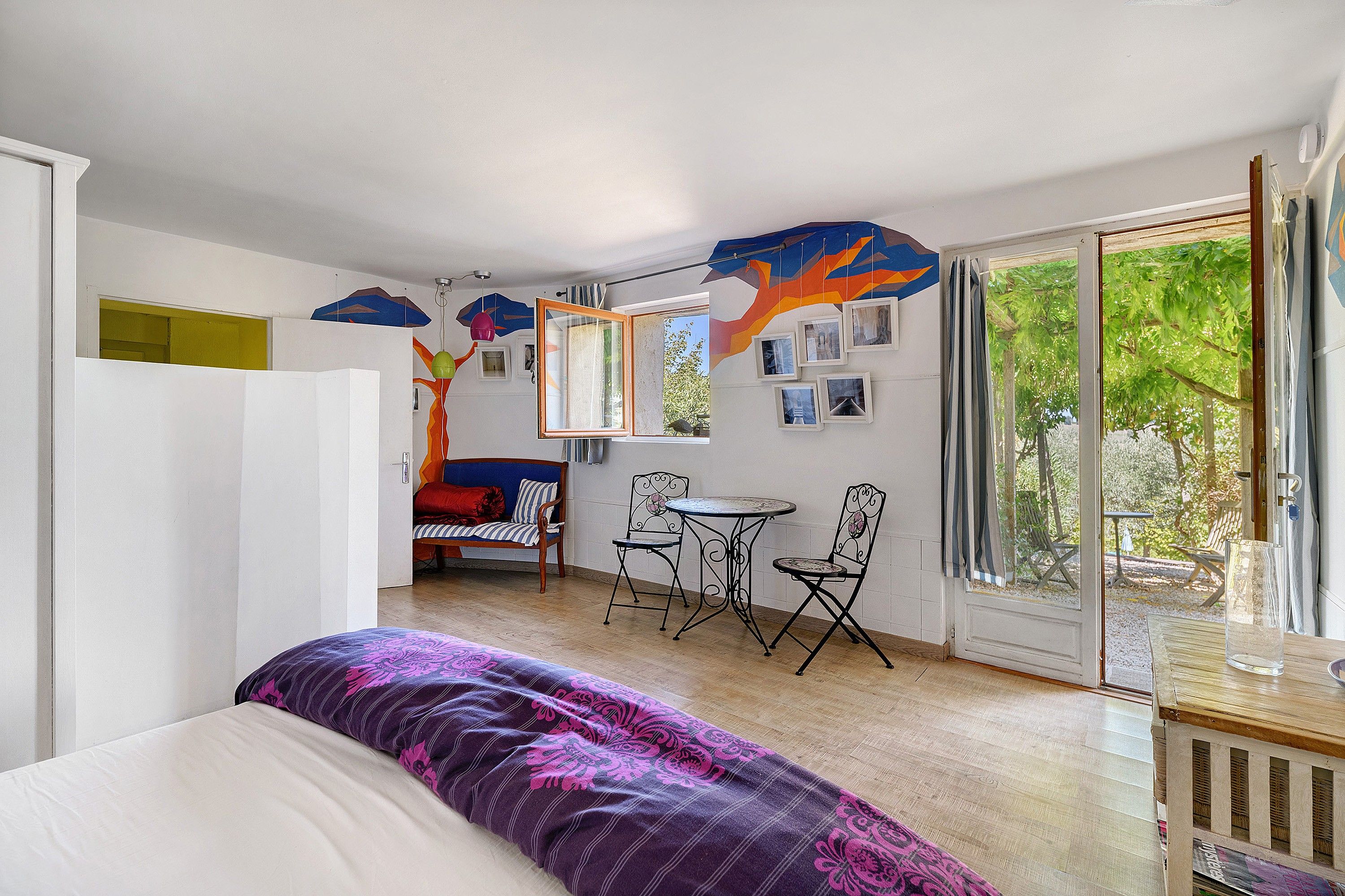 A room with a bed, a table with chairs, a sofa, and a large window with a view of greenery.