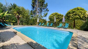 A rectangular swimming pool with a tiled edge, surrounded by lounge chairs and umbrellas, set against a backdrop of trees and greenery.