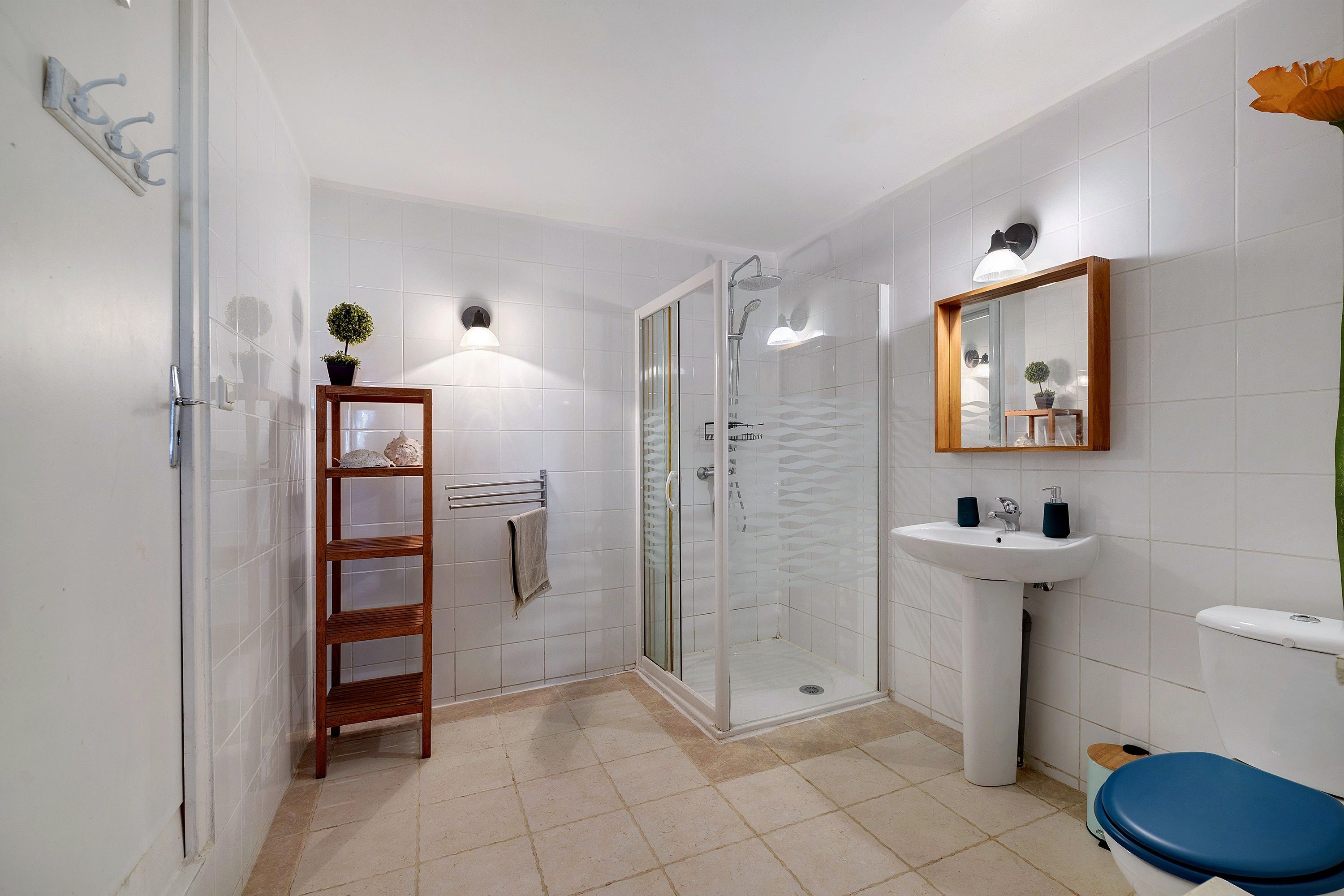 A bathroom with a glass shower enclosure, a wooden shelf with toiletries, a white sink, and a blue toilet.