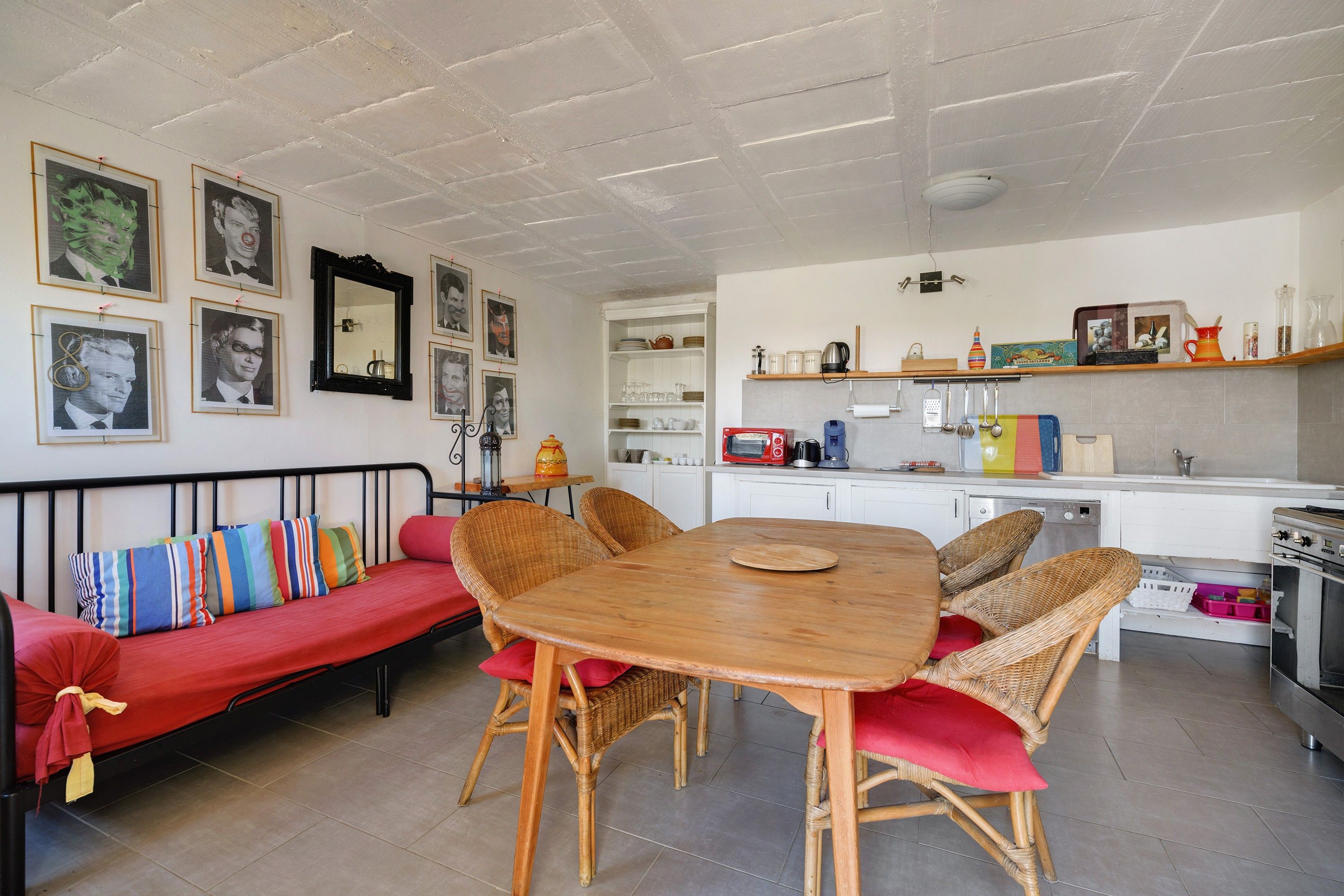 Un espacio de sala acogedor con una mesa de comedor de madera rodeada de sillas de mimbre, un sofá rojo con cojines de colores y una cocina al fondo.