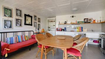 Un espacio de sala acogedor con una mesa de comedor de madera rodeada de sillas de mimbre, un sofá rojo con cojines de colores y una cocina al fondo.