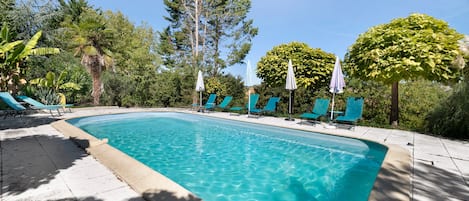 Une piscine rectangulaire à l’eau d’un bleu cristallin, entourée de chaises longues et de parasols, avec des arbres et de la verdure en arrière-plan.