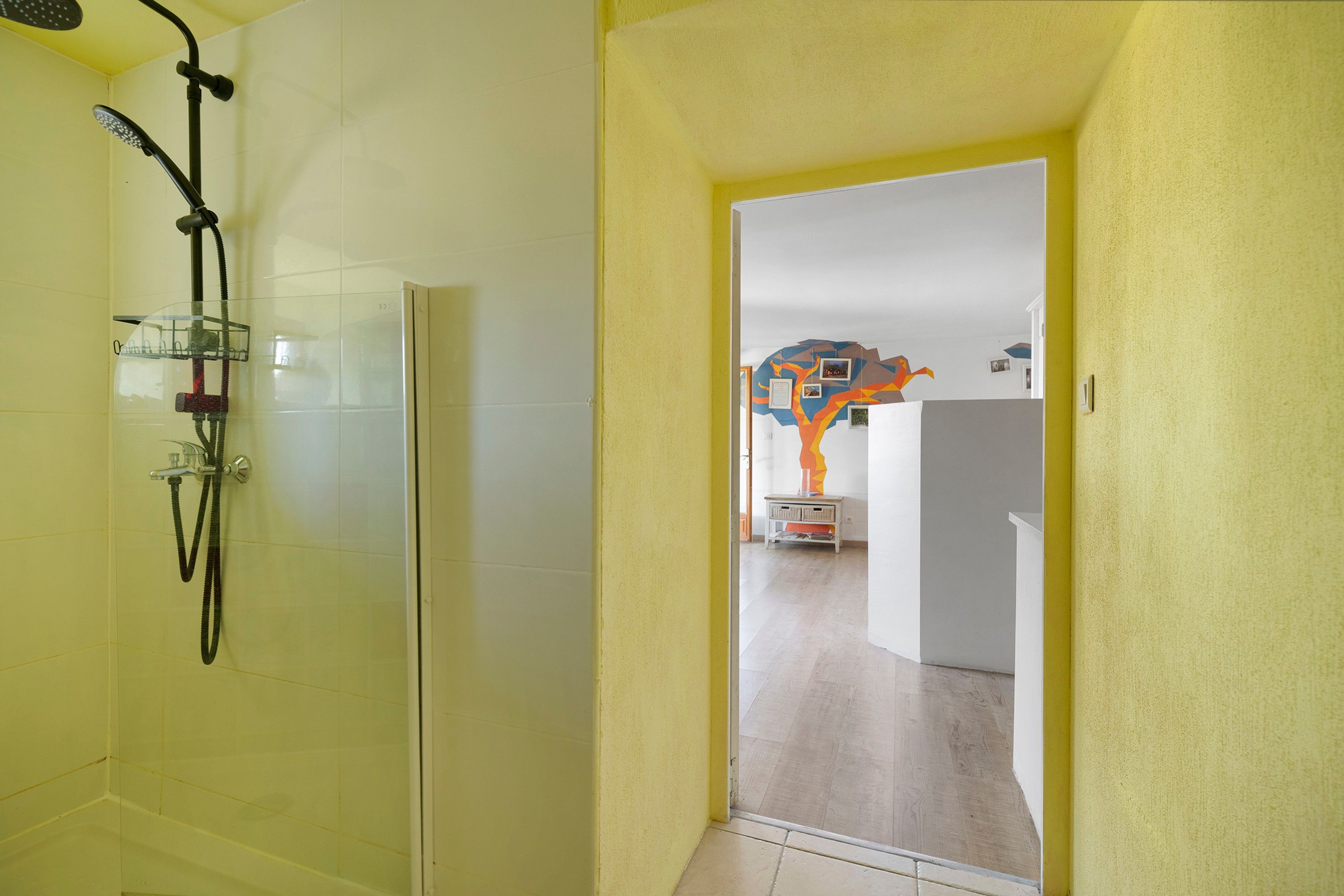 Une salle de bain avec une douche, des toilettes et un lavabo. Une porte donne accès à une autre pièce dont le mur est décoré de motifs colorés.