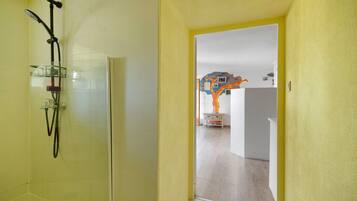 Une salle de bain avec une douche, des toilettes et un lavabo. Une porte donne accès à une autre pièce dont le mur est décoré de motifs colorés.